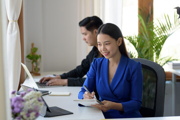 Asian businesswoman sitting happily with her laptop and takes notes intensely and smiles happily at her assignment.