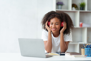 Cute young Asian woman wearing headphones listening to music happily on her laptop enjoying listening to work music during a relaxing break at work.