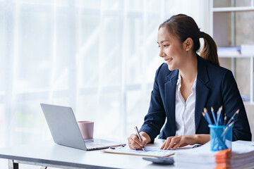 Asian businesswoman sitting happily with her laptop and takes notes intensely and smiles happily at her assignment.