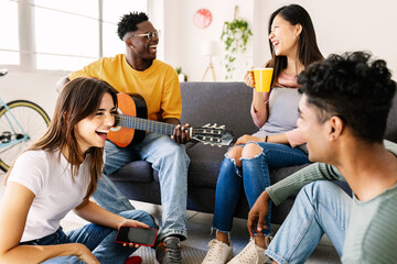 Young diverse friends having fun together at home - International group of teenage people enjoying free time together social gathering and playing the guitar while relaxing at apartment