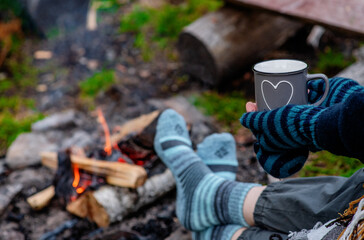 Traveller holds cup of the hot tea  and warms his feet by the fire