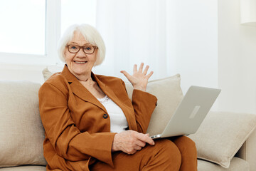 a very surprised elderly woman is sitting looking at a laptop on a sofa in a bright room near the window with glasses on her face and looks shocked with her hand raised up