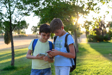 Two children students with their backpacks looking at their cell phones in a park.