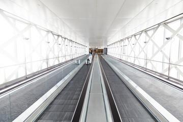 Black-haired Caucasian woman with a black backpack and a carry on walks alone on the airport conveyor belt.