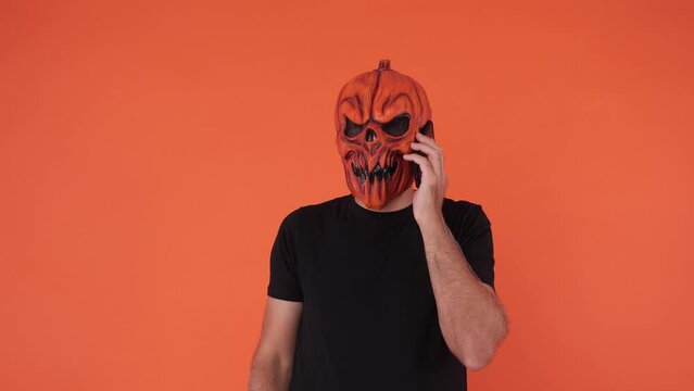 Person with pumpkin mask celebrating Halloween, looking at his mobile phone, on orange background. Concept of celebration, All Souls' Day and All Saints' Day.	