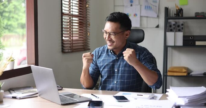 happy asian businessman and showing winner gesture while sitting at workplace in office.
