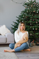 Lovely woman is sitting on the floor near the Christmas tree in the living room and holding a soft pillow in her hands and smiling