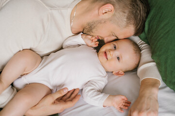 A handsome young dad lies in bed in the bedroom and hugs his loving laughing six-month-old boy. The concept of unconditional love and fatherhood