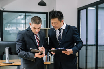 Diverse colleagues using tablet together, discussing online project, smiling asian businesswoman and businessman wearing glasses looking at device screen, standing in office
