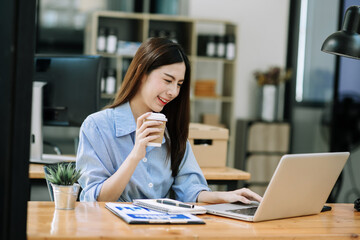 Fototapeta premium Confident beautiful Asian businesswoman typing laptop computer and digital tablet while holding coffee at office