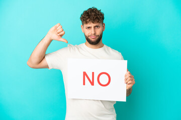 Young handsome caucasian man isolated on blue background holding a placard with text NO and doing...
