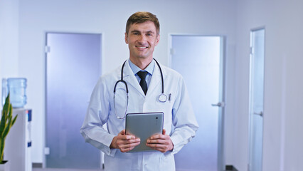 Modern hospital corridor portrait of a doctor man with a digital tablet looking straight to the camera and smiling large