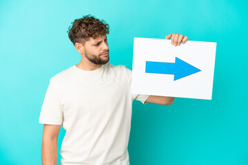 Young handsome caucasian man isolated on blue background holding a placard with arrow symbol