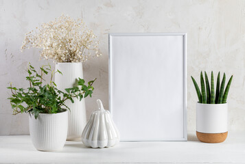 White trendy interior accessories. Frame, artistic pumpkin, houseplants, dry flowers, wooden table.