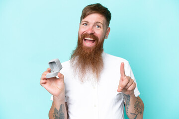 Redhead man with long beard holding a engagement ring isolated on blue background pointing up a...