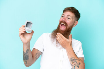 Redhead man with long beard holding a engagement ring isolated on blue background with surprise and...