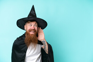 Young wizard in halloween isolated on blue background listening to something by putting hand on the ear