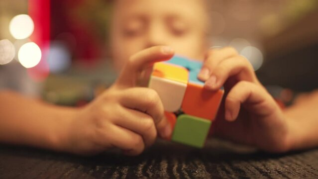 Close-up of hands child who collects rubik's cube of four colors on table. In blurred background face of caucasian baby boy and indoors restaurant. Games develop brain and knowledge. Puzzle for kid.
