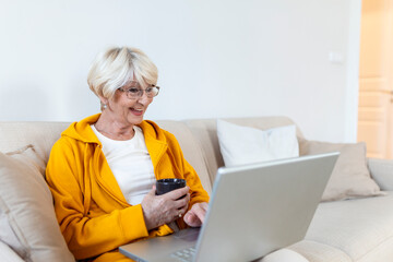 Older mature woman using wireless laptop apps browsing internet sit on sofa. Smiling middle aged woman having video call with her friends or family. Senior woman holding cup of coffee or tea