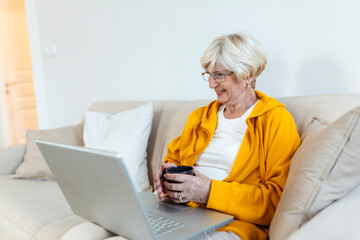 Fototapeta premium Enthusiastic progressive senior woman waving her hand and feeling happy while using a video call on laptop and talking with grandchildren. Elderly woman sitting on the sofa,holding cup of coffee