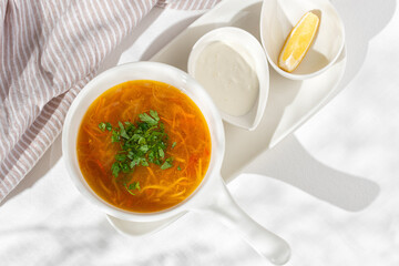 Fresh chicken soup with vegetables and pasta in a bowl with carrot and parsley on white table, top view