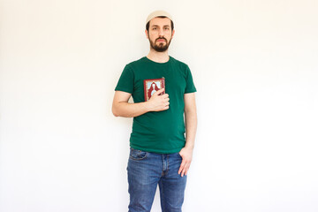Fototapeta premium Young bearded muslim man wearing a taqiyah (cap) and holding a Holy Quran, isolated on white background. Modern muslim guy wearing green t shirt and jeans.