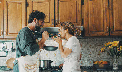 One happy real couple at home enjoying life together and relationship. Man giving food to woman to taste. Male people cooking for his wife. Male and female enjoying leisure activity indoor in kitchen - obrazy, fototapety, plakaty