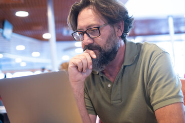 Portrait of man working on laptop in alternative store office. People and modern technology computer job leisure activity. Mature with beard and glasses using notebook at the table. Airport gate back