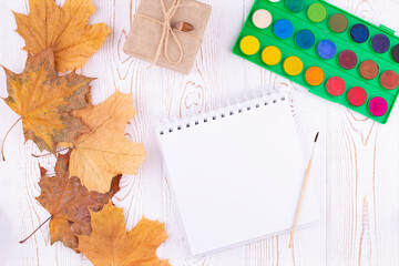 Autumn composition with empty sketchbook with spring, watercolor palette, autumn dry leaves, gift on wooden white background. Flat lay, copy space