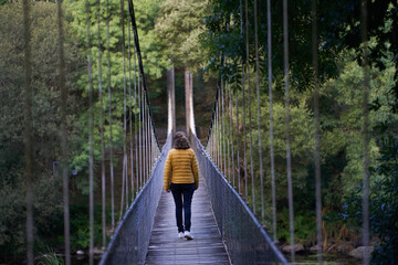 Close view of a woman with a yellow anorak walking on the suspension bridge with metal fence over the Miño river in Spain in an environment of native trees. 