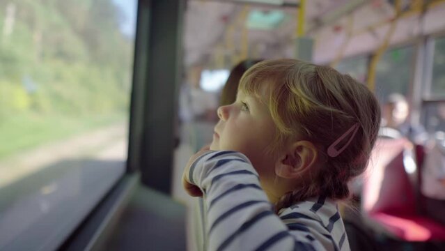 Little blonde girl enjoys the scenery from the bus window. The kid rides in public transport. The child looks dreamily through the glass. Thoughtful kids. Forest in the background.