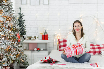 Beautiful brunette girl in a white sweater posing against the background of Christmas lights with a gift in her hands. Girl smiling and looking at the camera