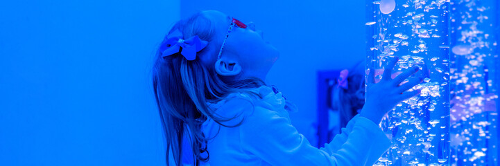 Child in sensory room, snoezelen, interacting with colored lights bubble tube lamp during therapy session. Child with cerebral palsy in occupational therapy. - obrazy, fototapety, plakaty