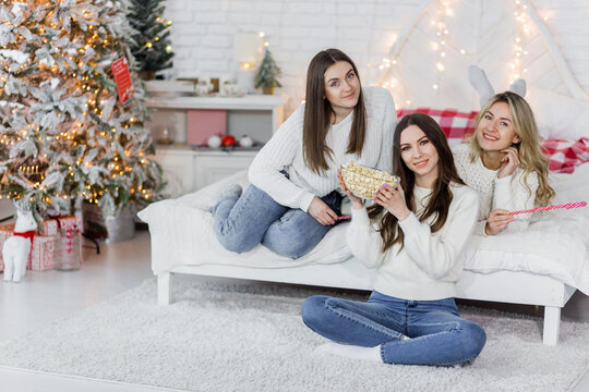 Group of young girl friends sitting on the floor and bed next to a Christmas tree, eating popcorn and looking to the camera.
