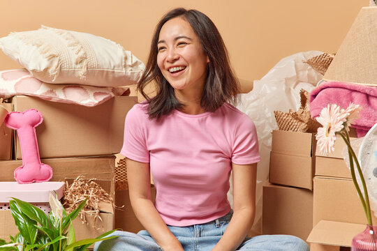 Overjoyed Asian Woman Laughs Out Gladfully Dressed In Casual T Shirt Focused Aside Surrounded By Carton Boxes Stacks Of Personal Belongings Relocates To New Apartment Isolated Over Beige Background.