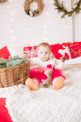 Obraz na płótnie Canvas A little girl under one year old in an airy dress on a large bed in a room decorated for Christmas, among the pillows of garlands and pine needles. Christmas mood. Children and Christmas