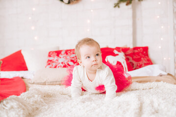 Fototapeta premium A little girl under one year old in an airy dress on a large bed in a room decorated for Christmas, among the pillows of garlands and pine needles. Christmas mood. Children and Christmas