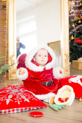 A little girl under one year old dressed as Santa Claus in a room decorated for Christmas, near the Christmas tree among the pillows, gifts, garlands and pine needles. Christmas mood. Children and Chr