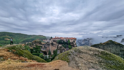 Fototapeta na wymiar Man standing on cliff edge with panoramic view of Holy Monastery of Varlaam, Kalambaka, Meteora, Thessaly, Greece, Europe. Rock formations overgrown with moss, moody atmosphere. UNESCO World Heritage
