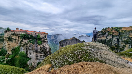 Fototapeta na wymiar Man standing on cliff edge with panoramic view of Holy Monastery of Varlaam, Kalambaka, Meteora, Thessaly, Greece, Europe. Rock formations overgrown with moss, moody atmosphere. UNESCO World Heritage