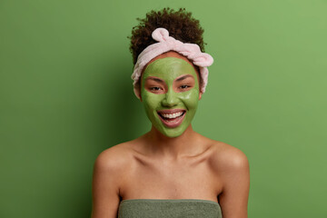 Happy curly haired woman wears headband wrapped in towel applies nourishing clay mask for skin treatment undergoes beauty procedures isolated over vivid green background. Rejuvenation and spa