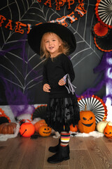 Smiling baby girl dressed as a witch with a magic wand in her hands for Halloween. Happy Halloween! Child in a witch costume with pumpkins in the dark