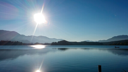 A panoramic view on the Faaker lake in Austrian Alps. The lake is surrounded by high mountains. Calm surface of the lake reflects the surrounding. Sweet flag at the shore. Clear blue sky. Serenity