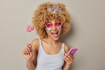 Funny woman with curly bushy hair holds caramel candy on stick and modern smartphone exclaims from joy applies silver patches under eyes wear small crown t shirt isolated over grey background