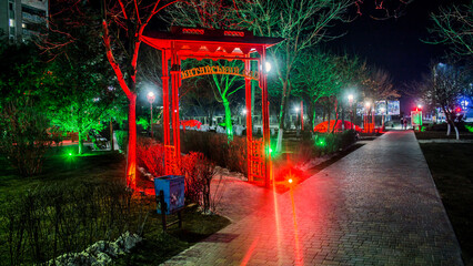 Chinese style red arch at night in front of the park with green and red lanterns and plants and stone path