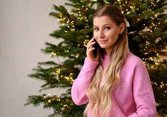 A beautiful girl in a pink sweater stands against the background of a Christmas tree in a cozy home interior and talks on the phone. The girl looks at the camera and smiles