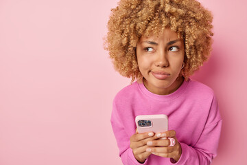 Serious confused woman holds mobile phone sends text messages dressed in casual jumper looks...