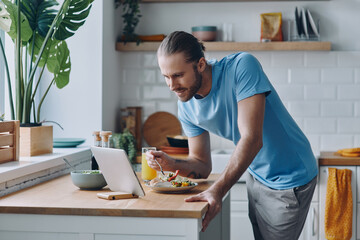 Handsome man eating lunch and looking at the digital tablet while standing at the kitchen counter