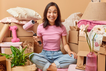 Happy Asian woman holds paint roller to improve apartment relocates to new flat surrounded by cardboard boxes full of household items against beige background. People real estate and mortgage concept