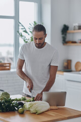 Fototapeta premium Handsome young man cutting veggies while standing at the kitchen island at home
